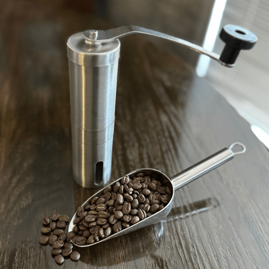 A manual stainless steel coffee grinder with a crank handle stands on a dark wooden table beside a metal scoop filled with roasted coffee beans. Coffee Grinder - Manual - Passport Coffee & Tea - Shop