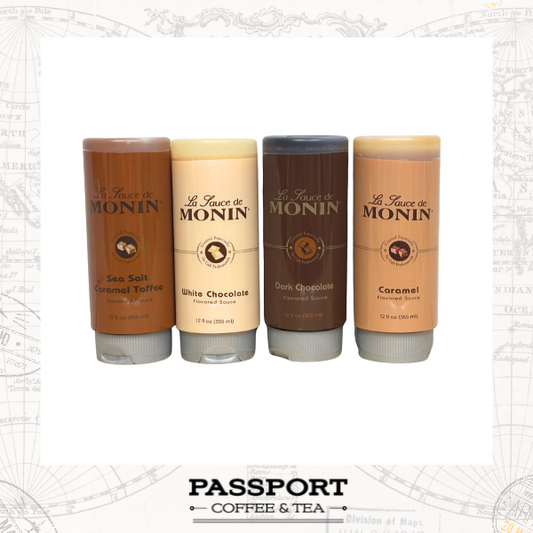 Four tubes of La Sauce de Monin coffee and tea flavoring sticks arranged in a row on a white background with a faint map border. The flavors shown are Sea Salt Caramel Toffee, White Chocolate, Caramel, and Dark Chocolate. 12oz bottles