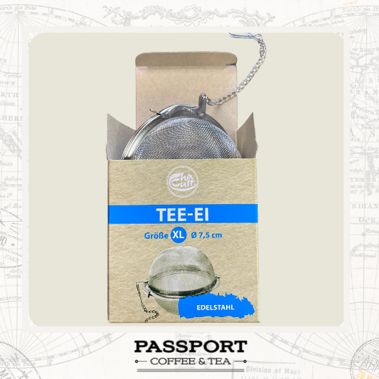 A metal tea infuser strainer with a hanging chain, shown partially inside its kraft paper packaging box. The box has a blue band reading 'TEE-EI' and 'Grösse XL Ø 7,5 cm', with a blue tab labeled 'EDELSTAHL'. With Passport Coffee & Tea label at the bottom.